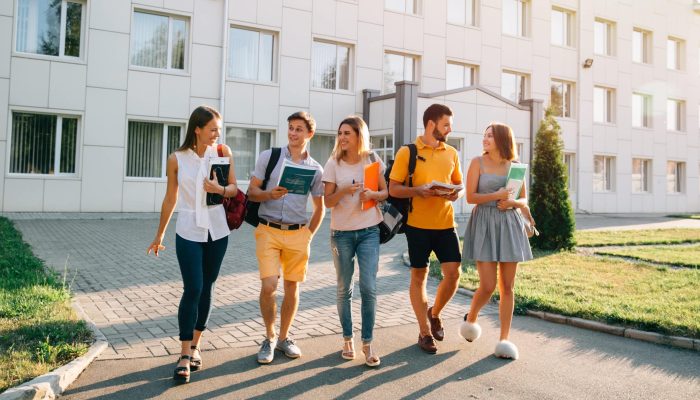 Free time of a students, bachelor`s campus life rhythm. Five friendly students are walking after they passed test outside the college building and discuss the project, smiling, enjoying, carefree